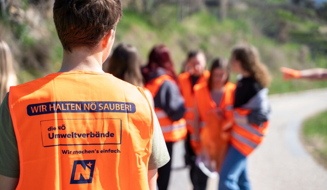 Ein Bub steht mit dem R&uuml;cken zum Fotograf, er tr&auml;gt eine Fr&uuml;hjahrsputz-Warnweste und im Hintergrund sammeln andere Jugendliche Abfall vom Boden auf