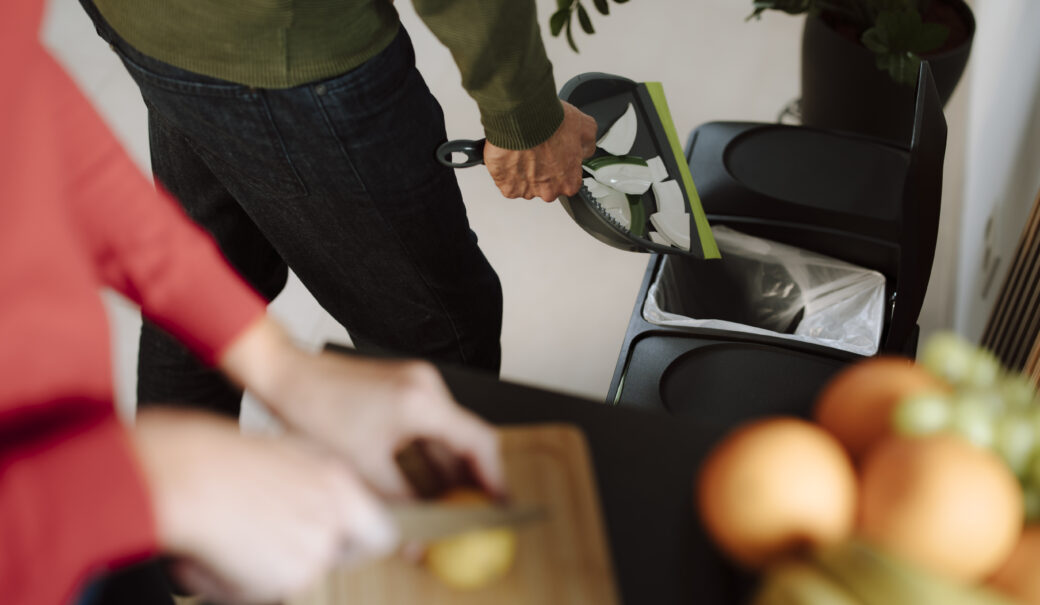 n der K&uuml;che wird besch&auml;digtes Geschirr entsorgt, w&auml;hrend im Vordergrund eine Zwiebel geschnitten wird.