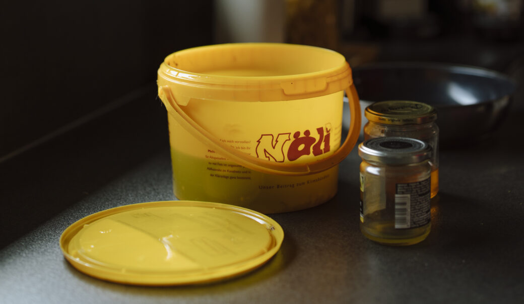 Auf dem Bild ist ein N&Ouml;LI Beh&auml;lter zu sehen. Davor liegt der Deckel und rechts davon stehen zwei Gl&auml;ser mit Metall-Schaubdeckel, die mit &Ouml;l gef&uuml;llt sind.