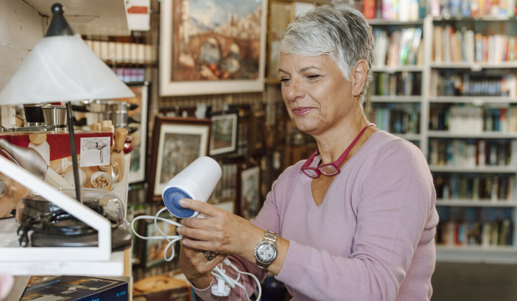 Auf dem Bild ist eine Frau mittleren Alters mit kurzen, grauen Haaren zu sehen. Sie tr&auml;gt einen hellvioletten Pullover und eine silberne Armbanduhr. Um ihren Hals h&auml;ngen rote Lesebrillen. Die Frau h&auml;lt ein kleines elektrisches Ger&auml;t mit Kabel in den H&auml;nden, vermutlich pr&uuml;ft sie es oder betrachtet es genauer. Sie befindet sich in einem Second-Hand- oder ReUse-Laden, umgeben von Regalen voller unterschiedlichster Gegenst&auml;nde: K&uuml;chenger&auml;te, B&uuml;cher, Bilderrahmen, Lampen und Spielzeug. Im Vordergrund ist ein pinkes Pl&uuml;schspielzeug erkennbar. Der Hintergrund zeigt weitere Regale mit B&uuml;chern und Haushaltsartikeln, was den Eindruck eines gut sortierten, aber abwechslungsreichen Shops vermittelt. Das Bild strahlt eine ruhige, konzentrierte Stimmung aus.